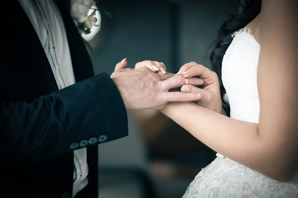 Woman putting ring on husbands hand in photo. White Gown and Black Tuxedo Wedding Insurance and Wedding Event Insurance.